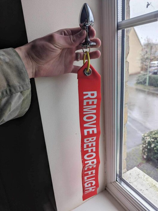 Hand håller upp en silverfärgad beslagshållare med en röd etikett som läser "REMOVE BEFORE FLIGHT" vid ett fönster.