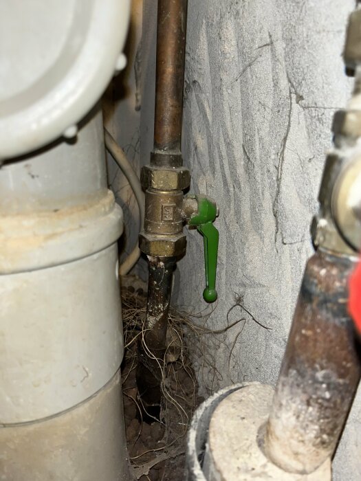 Vattenledningar med en grön och en rostfärgad spak i ett hus, omgivna av rör och kablage.