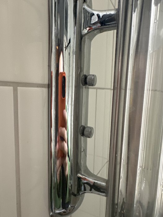 Närbild av duschväggsfäste mot kakelvägg, med två synliga skruvar och en reflekterad spegelbild.