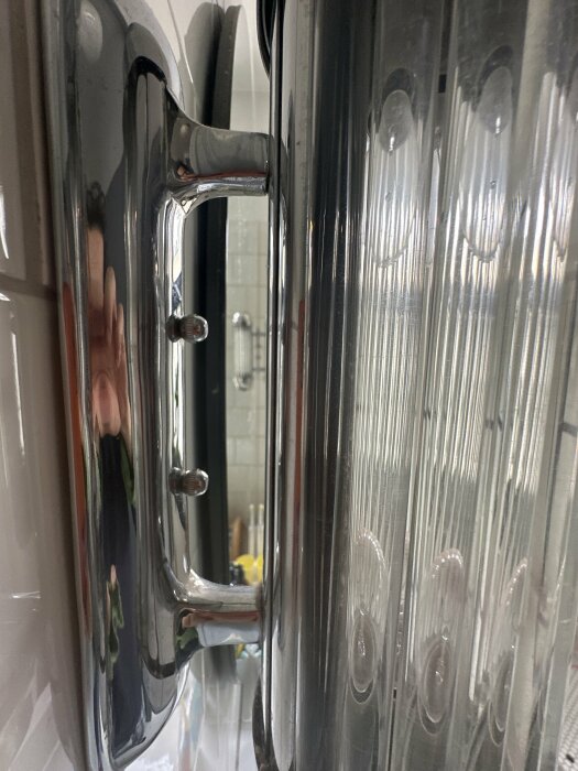 Närbild på en duschdörrs fäste mot väggen med reflekterande ytor och suddig bakgrund.