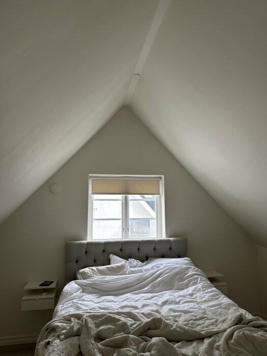 Sovrum med snedtak och fönster utan takbjälkar, vitmålade väggar och säng med täcke.