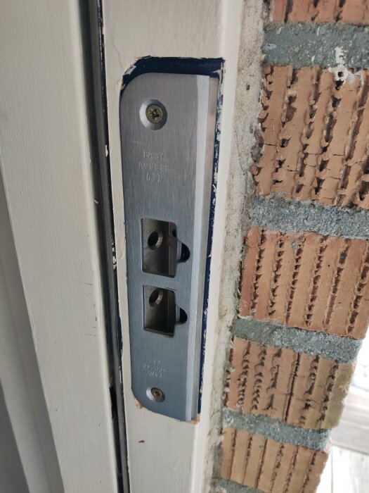 Ett låshus monterat på en vit dörrkarm nära ett tegelvägg, med synligt justering för tryckesfall.