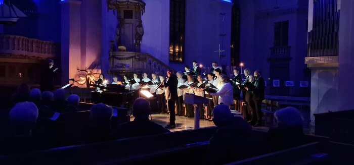 Kör framför publik i en kyrka med blå belysning och en pianist till vänster.
