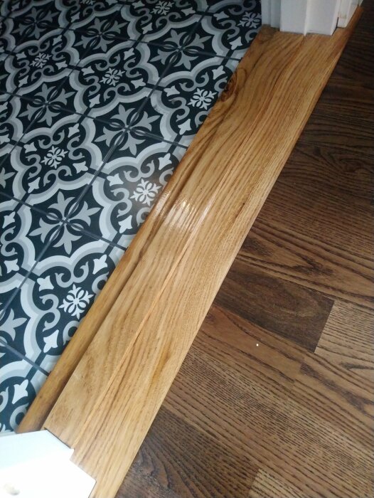 Handgjord tröskel av ek som ligger mellan två olika golv, ett mönstrat kakelgolv och ett mörkt trägolv.
