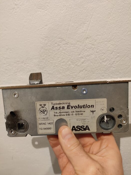 Hand håller en dörrlåsmekanism märkt "Assa Evolution" med synliga monteringsskruvar.