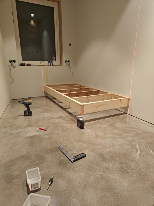 Träram för sängkonstruktion på ett betonggolv med verktyg och laserlinjer runt omkring.