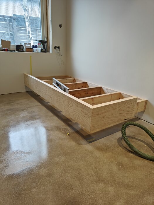 Sängstomme av askträ under konstruktion i ett rum med betonggolv och verktyg.