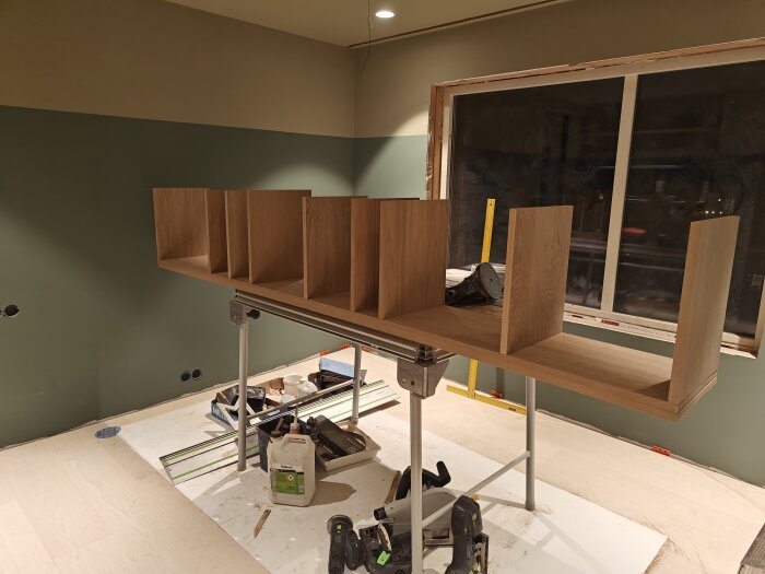 Pågående bygge av en trähylla som står på en arbetsbock i ett outfärdigt rum med verktyg.