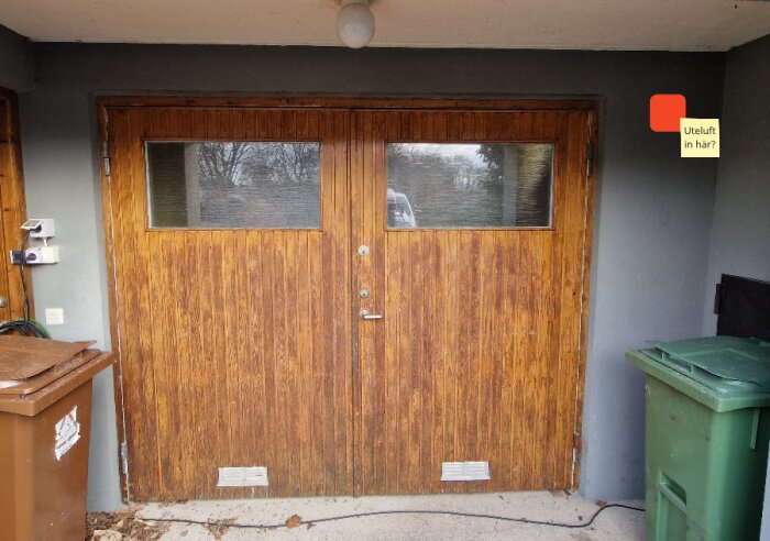 Träfärgad garageport med två fönster och en etikett markerad "Uteluft in här?" ovanpå på höger sida.