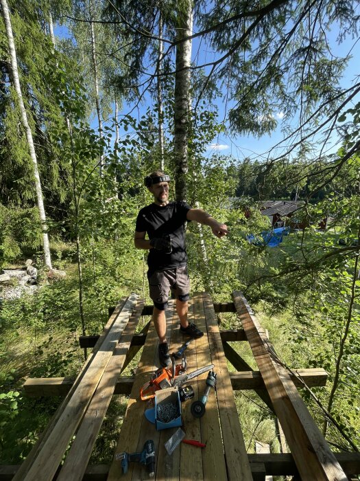 Person står på en träkojans plattform uppe i träd med verktyg och byggmaterial runtomkring.