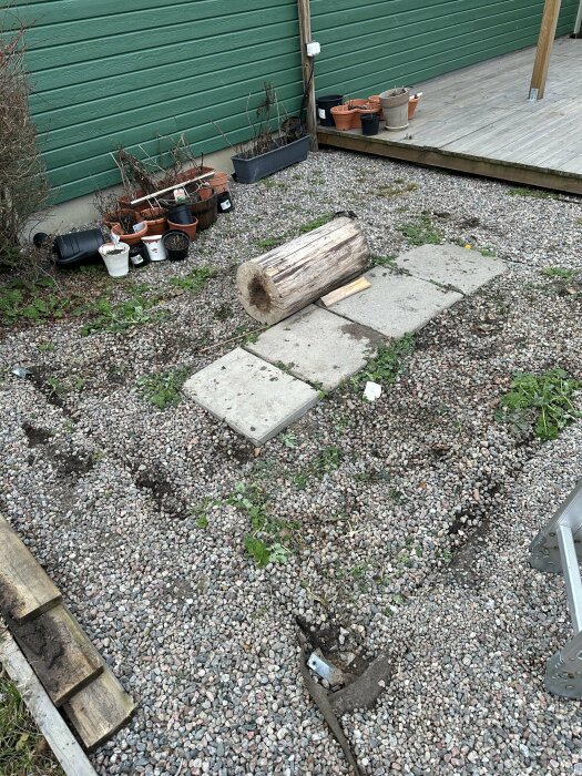 Gruslagd yta med spår och fundament efter ett borttaget växthus, krukor och en trädstam nära en träveranda.