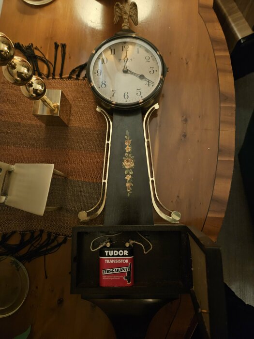 Antik pendylväggklocka med svart och guldinredning och Tudor Transistor batterigaranti skylt.
