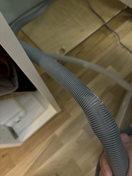 Klämd grå Aquastop+-slang till diskmaskin som hålls framför en diskmaskin i ett kök.