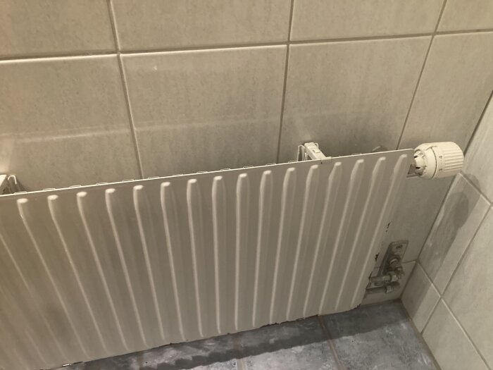 Vit radiator installerad under ett fönster med kakelvägg och grått golv.