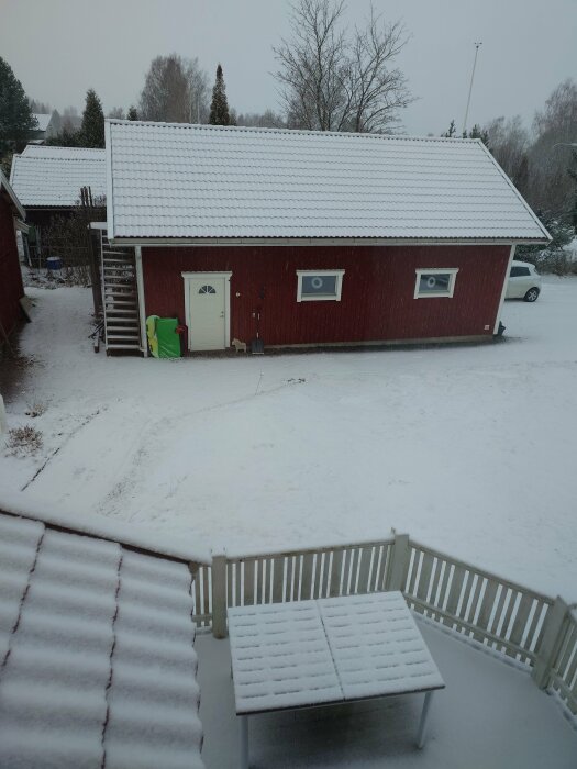 Ett rödmålat hus med snötäckt tak och omgivning, inklusive en vit veranda och bord.