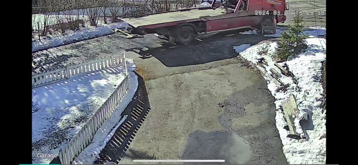 Övervakningsbild på en röd bärgningsbil vid en uppfart med snörester och staket.