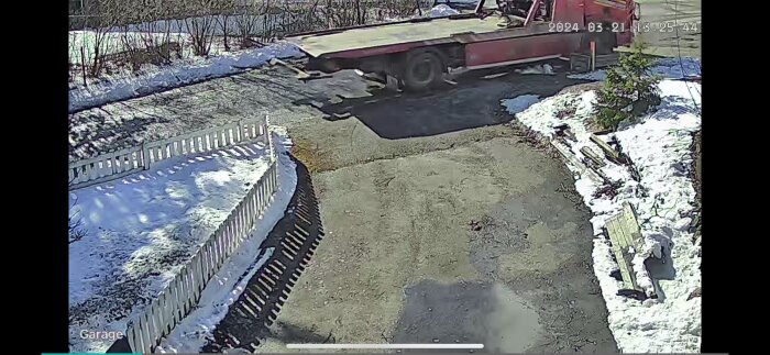 Övervakningsbild på en röd bärgningsbil framför en uppfart med snö och staket.