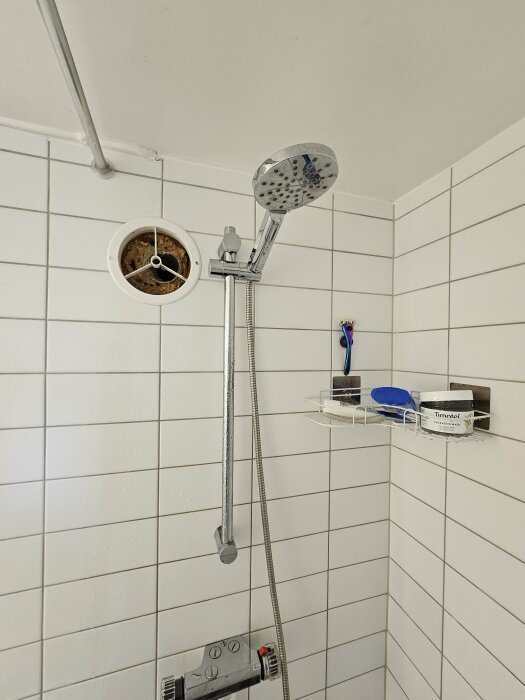 Bild på ett badrum med kaklade väggar, där en gammal frånluftskanal och en duschhylla med hygienartiklar syns.