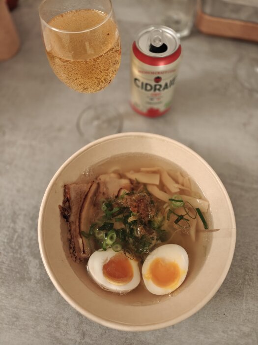 Bild på en skål ramen med ägg, gräslök och skivor av kött, intill en burk cidrar och ett glas med dryck.