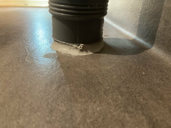 Silikon applicerat vid foten av ett svart handfat på en grå matta för att täta ett område nära golvet.