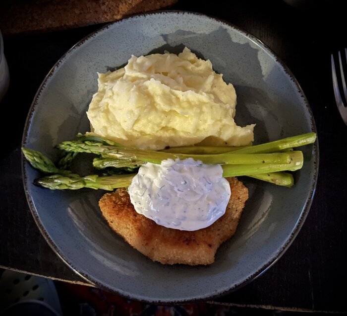 Panerad plattfisk med en klick dillsås, hög med potatismos och kokt grön sparris på en grå tallrik.