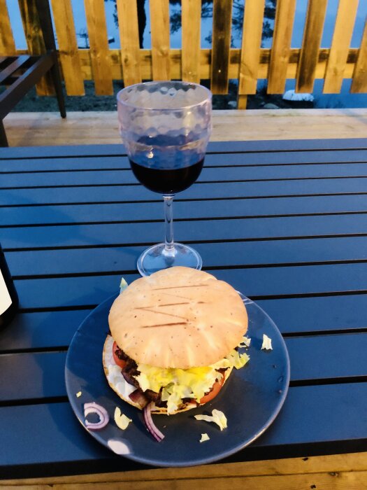 Grillad hamburgare på tallrik med rödlök, sallad och tomat, glas rödvin, på ett utomhusbord, trästaket i bakgrunden.