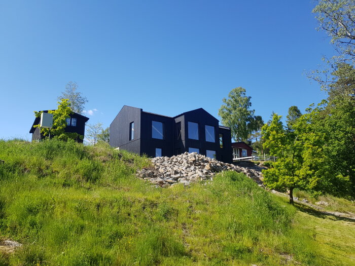 Modernt svart hus under konstruktion på en gräsbeklädd sluttning med klippor och träd under blå himmel.