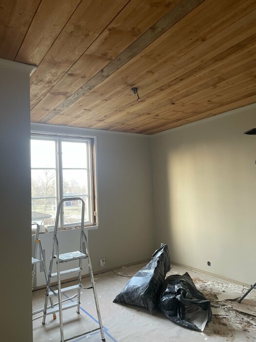 Renoverat rum med nytt träpaneltak, oslipat golv och byggstege nära ett fönster.