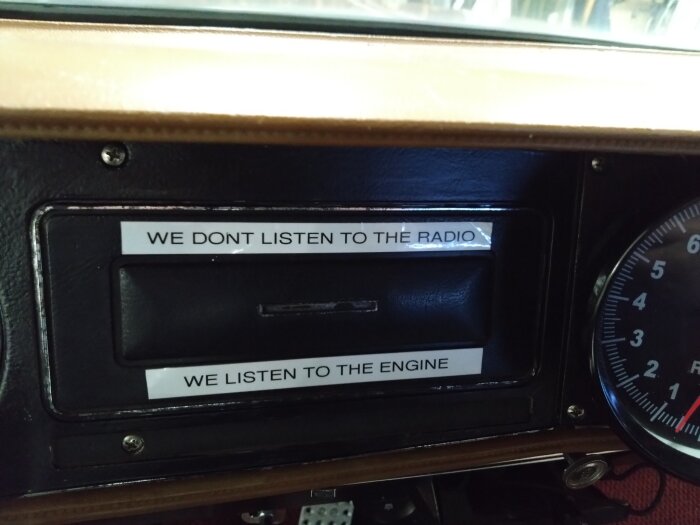 Instrumentbräda med skylt som säger "WE DONT LISTEN TO THE RADIO WE LISTEN TO THE ENGINE" bredvid varvräknare.