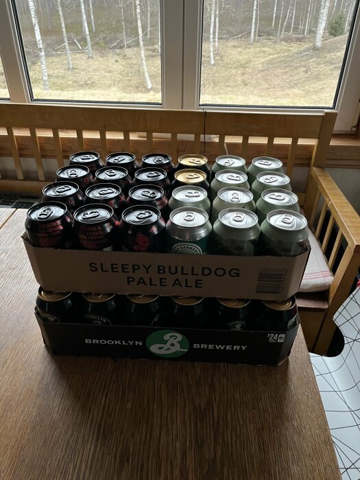 Flak med burkar av Sleepy Bulldog Pale Ale och Brooklyn lager på ett bord framför ett fönster med utsikt över skog.