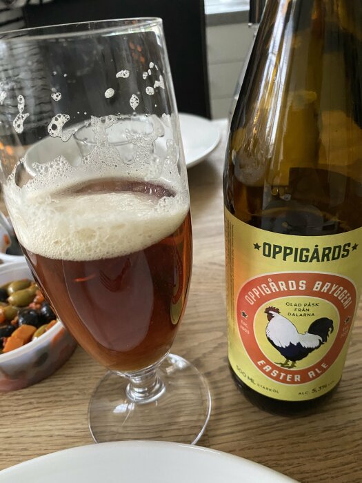 Ett glas med mörkt öl och en Oppigårds bryggeri Påsköl flaska på ett bord med delar av en måltid.