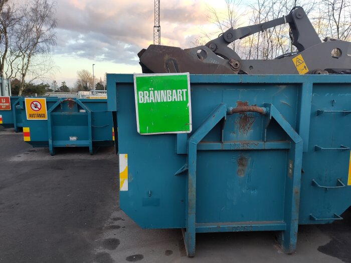 Blå container märkt BRÄNNBART med skylt för avstånd och grävmaskin i bakgrunden på ÅVC.