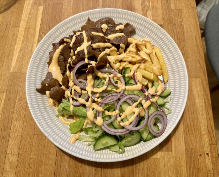 En välserverad kebabtallrik med kött, pommes frites, sallad, rödlök och sås på en vit tallrik.