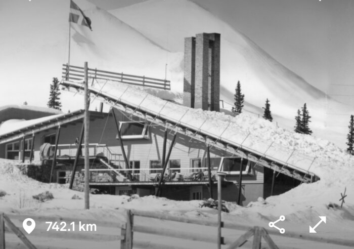 Svartvit bild av Erskine-designat byggnad i snölandskap med skidbacke och skorstensliknande struktur.