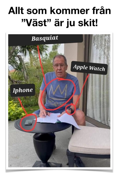 Man i t-shirt med text och symbol, har iPhone och Apple Watch, sitter vid ett bord med papper.