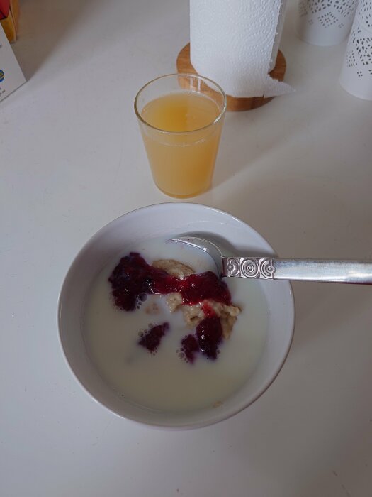 Skål med havregrynsgröt och sylt, bredvid ett glas juice och en pappersrulle på ett köksbord.