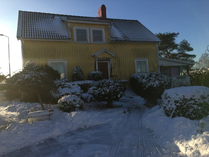 Ett tvåvåningshus med gult träfasad och snötäckt tak och trädgård i vintermiljö.