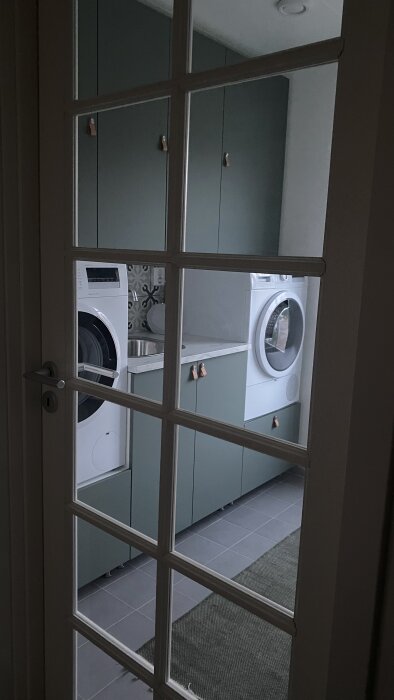 Tvättmaskin och torktumlare inbyggda under bänkskiva, sett genom glasvägg med fönsterkors.