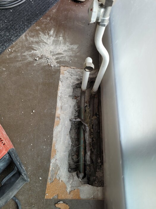 Uppgrävd golvsektion som visar två vattenrör nära en vit vägg och en del av en radiator.