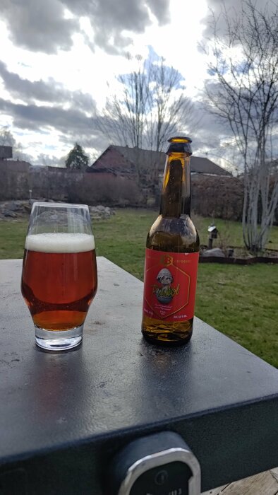 Ett glas med öl och en ölflaska utomhus på ett bord med trädgården i bakgrunden.