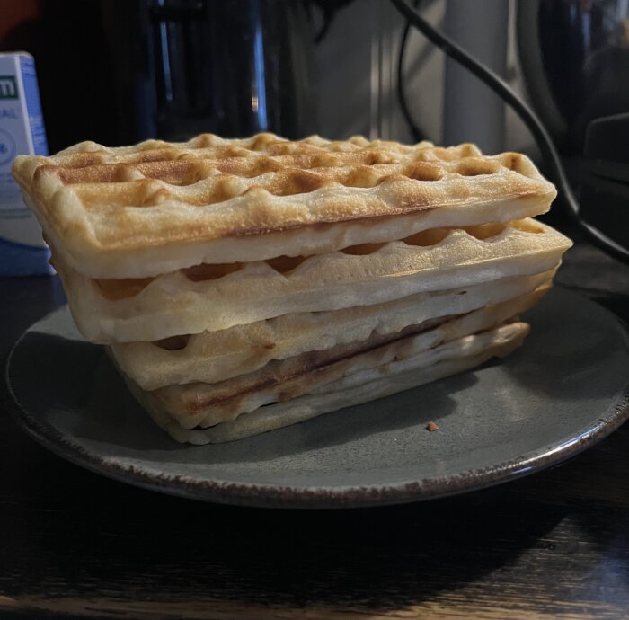 Fräscha våfflor staplade på en mörk tallrik med blåbär och grädde i bakgrunden.