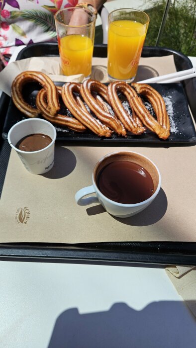Churros med pudersocker, kopp med drickchoklad, nutella och två glas apelsinjuice på ett cafébord.