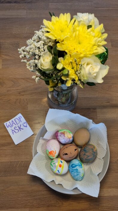 En bukett blommor bredvid en tallrik med målade påskägg, samt en lapp med texten "GLAD PÅSK".