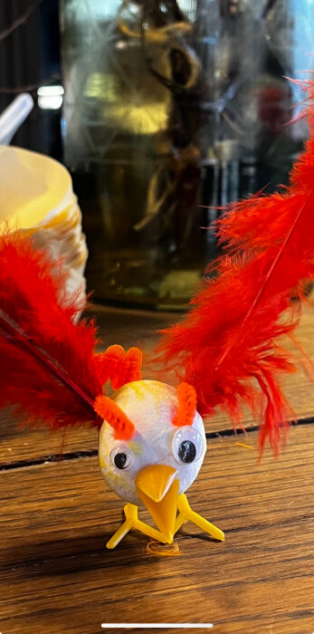 Handgjord påskkyckling med röda fjädrar och gula detaljer på ett träbord.