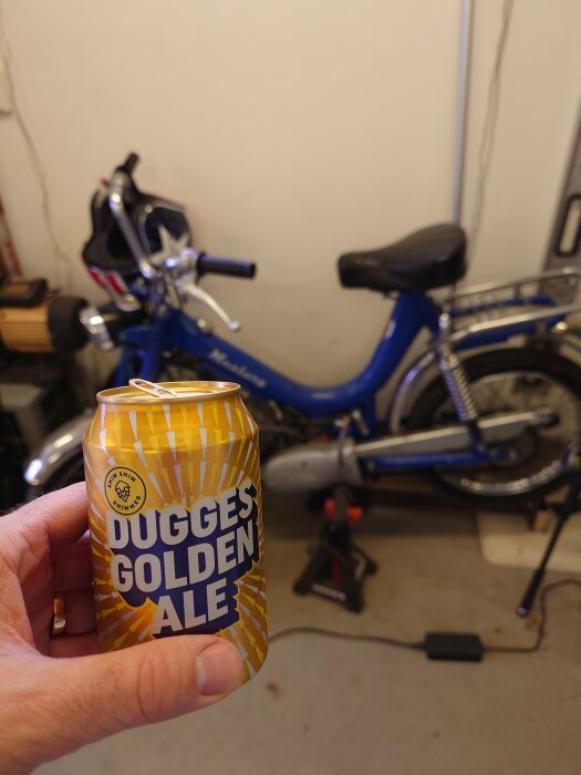 Hand håller en ölburk med texten "DUGGES GOLDEN ALE", suddig bakgrund med blå moped i ett garage.