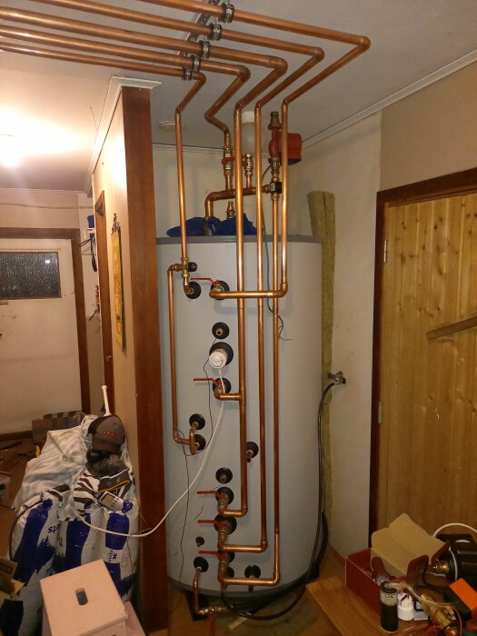Installation av en varmvattenberedare med kopparledningar och anslutningsdetaljer i ett rum med diverse byggmaterial.