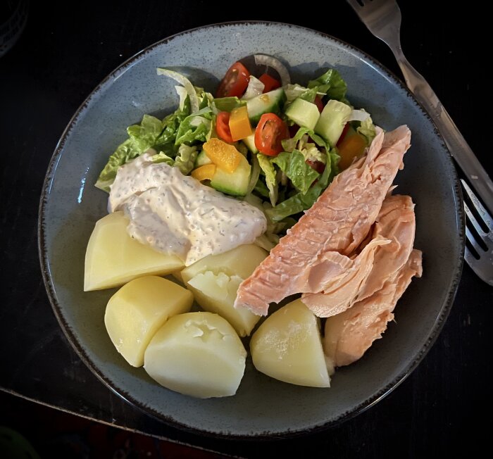 Tallrik med varmrökt lax, kokt potatis, sallad och romsås serverad på ett bord.