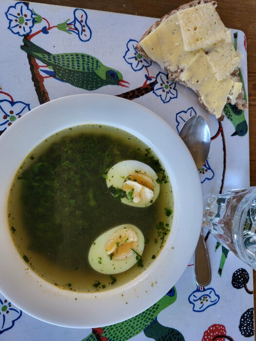 Nässelsoppa med halverade ägg i en vit skål, knäckebröd med Västerbottenost, på ett påskdukat bord.
