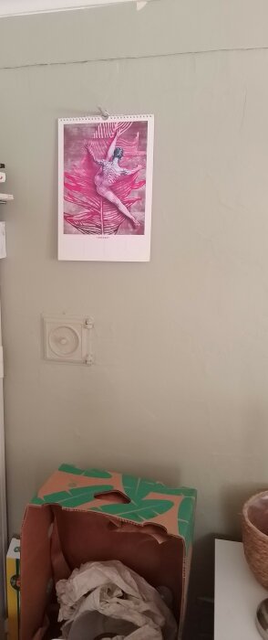 En almanacka med konstnärlig bild på en vägg ovanför en kartong och en vit ventil på en gråmålad vägg.