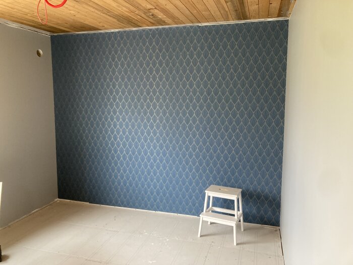 Nyrenoverat rum med nymålad vägg och en tapetserad vägg med blått mönster, liten vit pall på golvet.